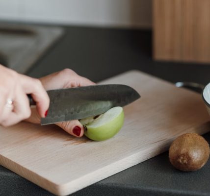Crop unrecognizable female cutting green apple with sharp knife while standing in light kitchen near counter with kiwi and bowl near cutting board