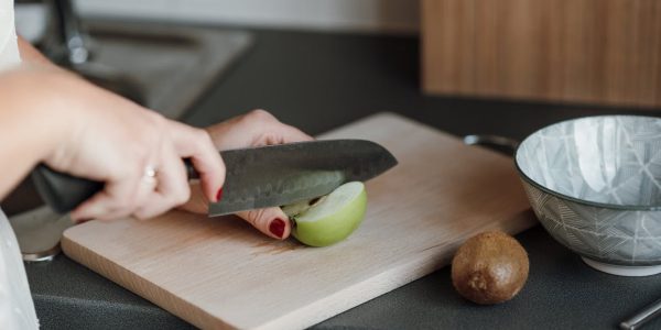 Crop unrecognizable female cutting green apple with sharp knife while standing in light kitchen near counter with kiwi and bowl near cutting board