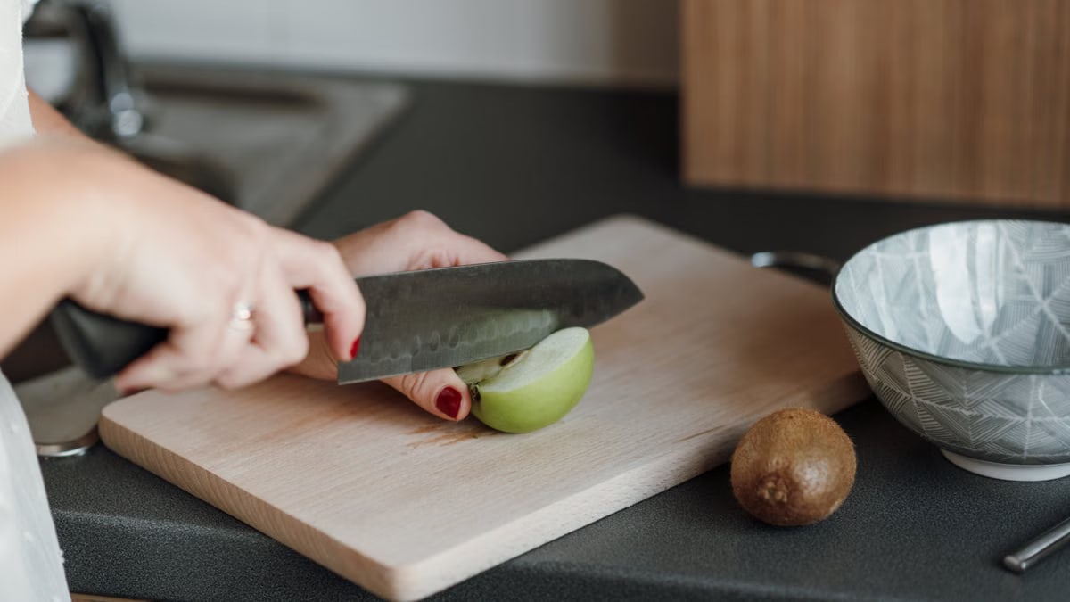 Crop unrecognizable female cutting green apple with sharp knife while standing in light kitchen near counter with kiwi and bowl near cutting board