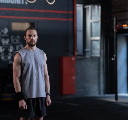 Muscular man standing in a gym with gymnastic rings, showcasing fitness and strength.